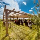 Group of people mingling under a wooden pergola with picnic tables at an outdoor event on a sunny day; a white fluffy dog nearby on the grass.