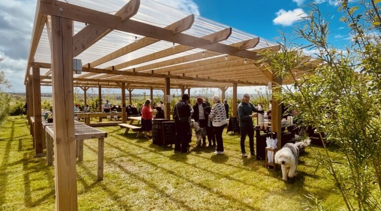 Group of people mingling under a wooden pergola with picnic tables at an outdoor event on a sunny day; a white fluffy dog nearby on the grass.