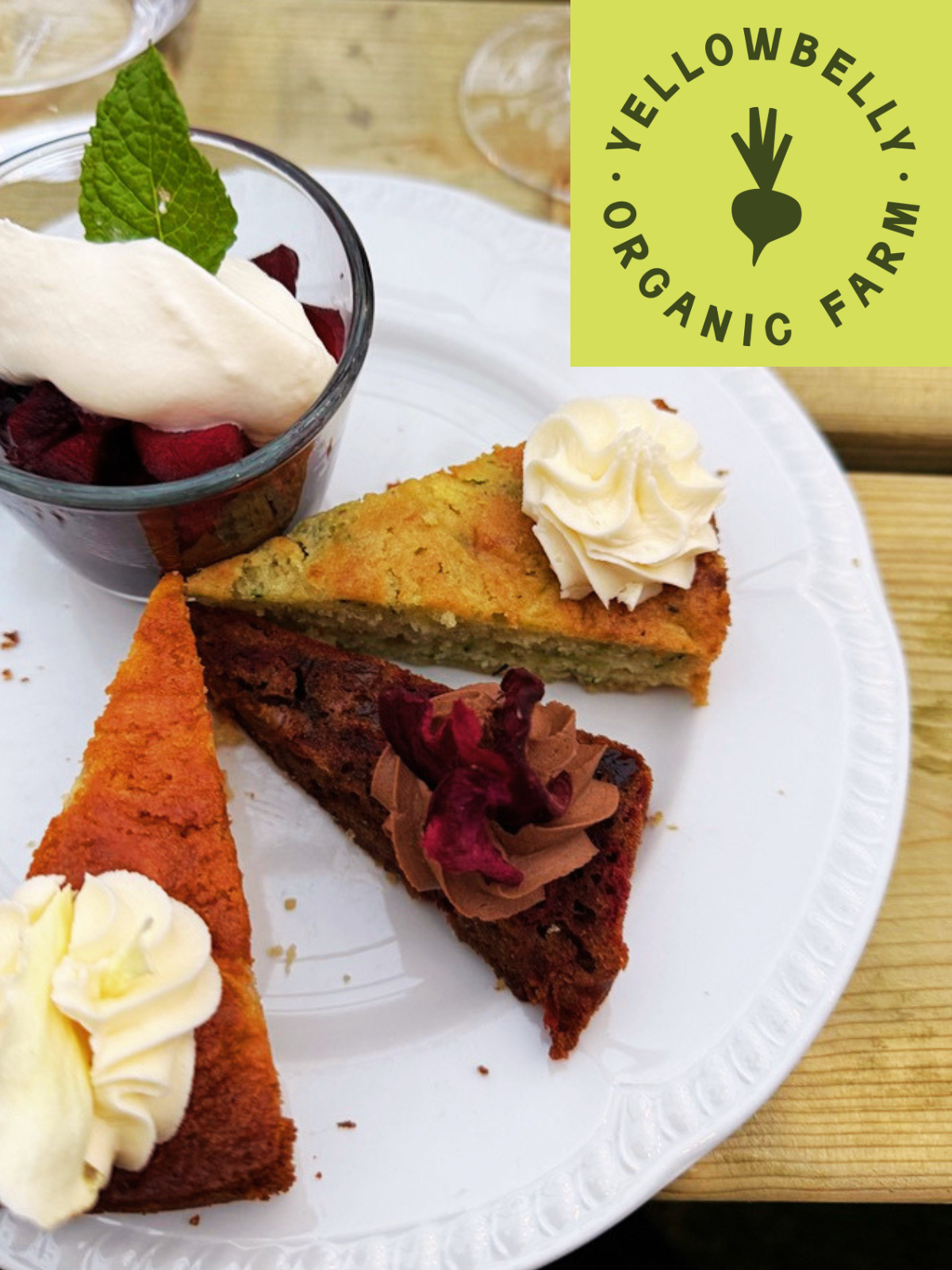 Plate of assorted cakes with whipped cream, mint leaf garnish, and rose petals on a white plate; Yellowbelly Organic Farm logo in the top-right corner.
